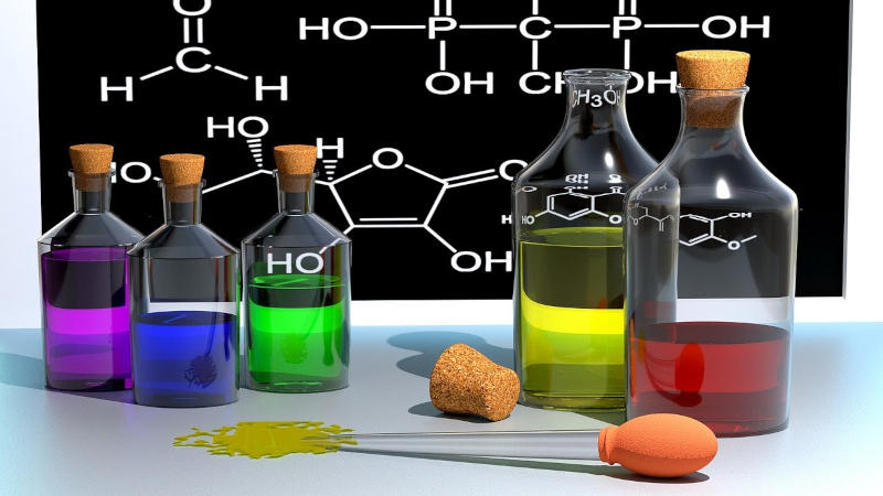 Flaschen mit farbigen Flüssigkeiten vor einer Tafel mit einer chemischen Formel