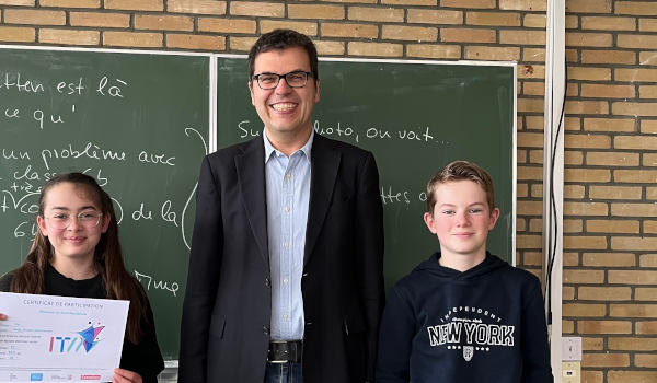 Zwei Schüler*innen und Schulleiter Dr. Flatten in einem Klassenraum; eine Schülerin präsentiert eine Urkunde.