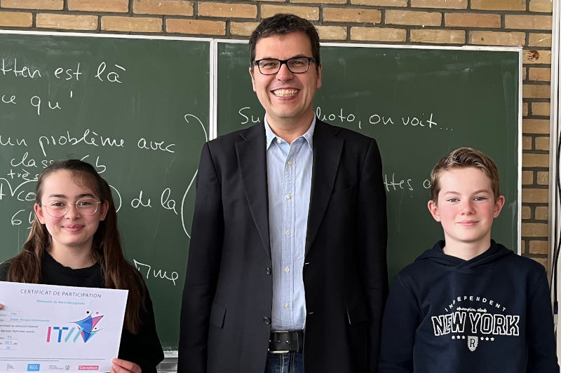 Zwei Schüler*innen und Schulleiter Dr. Flatten in einem Klassenraum; eine Schülerin präsentiert eine Urkunde.