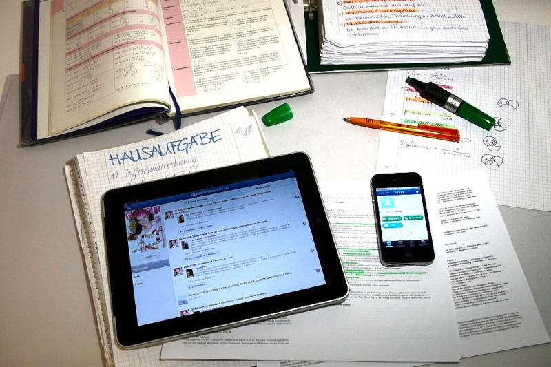 Ein Tisch, auf dem ein Block mit Überschrift "Hausaufgabe" liegt, außerdem Bücher, Kopien, Stifte, ein Tablet und ein Smartphone